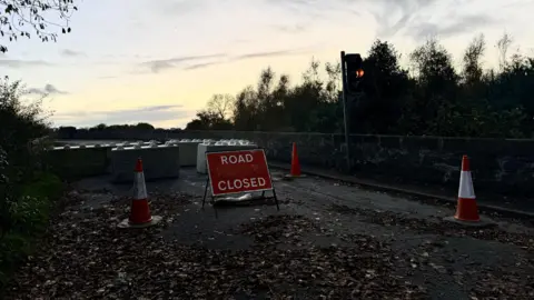 A road closed sign approaching Kilrea Bridge. Traffic cones and large blocks have been placed across the road. A single traffic light shows an amber signal.