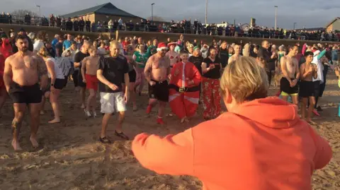 Alan Hughes Dozens of people on the sand at Berwick, many in shorts. Some are wearing Santa hats as they prepare to run into the sea. 