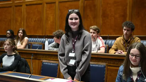 Cara smiles from the seats in the assembly chamber. 