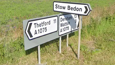 A road sign at a junction on the A1075 showing directions for Thetford, Dereham and Watton. An arrow points to a side road, showing the direction for Stow Bedon.