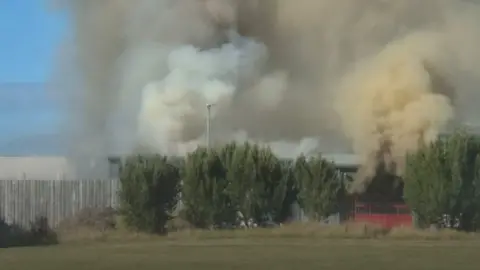 Image shows smoke pouring from a fire station on the Isle of Man. Thick white and brown smoke is rising into the sky behind a line of trees. 