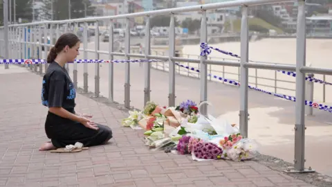 BBC/Isabelle Rodd Katherine Pierce kneels in front of floral tributes and prays