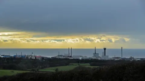 A general view of the Sellafield site pictured from afar at sunset. Green fields are visible in the foreground, with the industrial buildings, including chimneys and the sea in the background.