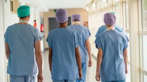 Getty Images Five nurses walking down a hospital corridor. Their backs are turned to the camera and they are wearing blue scrubs and surgical hair nets. 