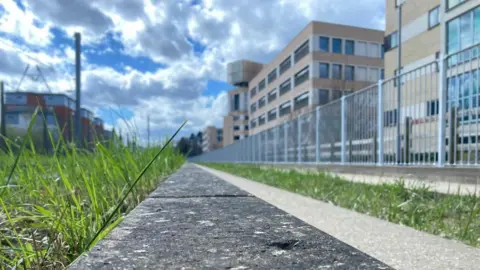 Jozef Hall/BBC A strip of grey concrete bordered by green grass. Building line either side of the strip. On the right-hand side in a grey metal fence. In the distance is a cloudy sky.