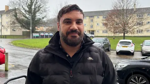 A man in his 40s with short brown hair swept to the side and dark facial hair smiles as he stands in the rain on a housing estate. Behind him is a road with cars parked on it, a large grass verge and a three-bed block of light coloured flats.