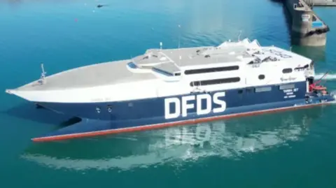 The picture shows a large high-speed ferry on calm blue water near a dock. The vessel has a sleek, modern design with a pointed bow and a flat upper deck. It is painted mainly white on the upper section and dark blue on the lower section, with the letters DFDS prominently displayed in bold white font along the side. 
