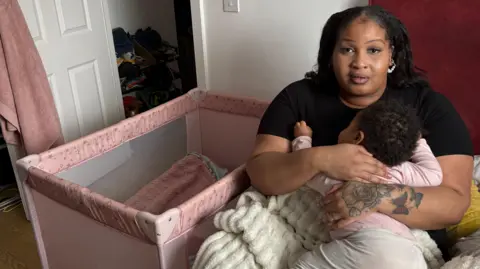 Martin Heath/BBC Laura-May with medium-length dark hair and a white string-of-pearls earring visible on her left ear. She is wearing a dark-coloured T-shirt and holding a young child who has black hair and is wearing a pink babygrow. She is sitting on her bed and has a light-coloured blanket over her legs. There is a pink cot to her right and a white door beyond that.