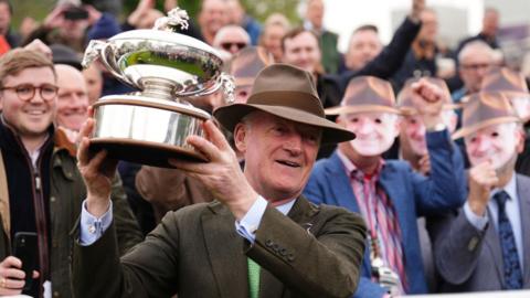 Willie Mullins with the champion trainers' trophy at Sandown, in front of fans wearing Willie Mullins masks