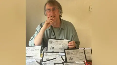 Andy Taylor A person sits at a table indoors, holding an old paper document while several similar documents are spread out in front. The papers appear to be historical banknotes or certificates, arranged neatly across the surface. The background is plain and softly lit, drawing attention to the documents and the person’s presence behind them.