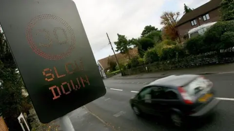 PA Media A black sign with lights reading 30 slow down at the side of a road. A slightly blurred blue car is on the road beneath the sign. A hedge in front of residential buildings on the opposite side of the street.