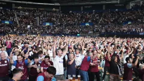 Reuters Claret and blue clad fans jump for joy in the arena. Most are in Villa tops. Many have their hands in the air at ground levels and in surrounding seating.