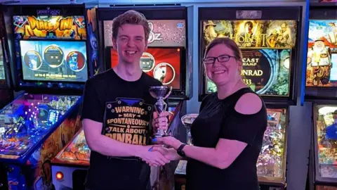 Pinfest office Nathan Garwood, wearing a black T-shirt, is presented with a trophy by a woman wearing a black top. There are pinball machines behind.