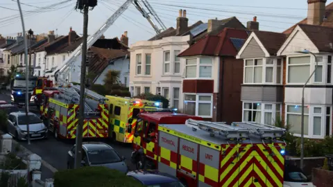 Kevin Boorman Several fire service appliances stationed at a residential road. There are vehicles parked on each side of the street. There is also a collapsing roof on a building.