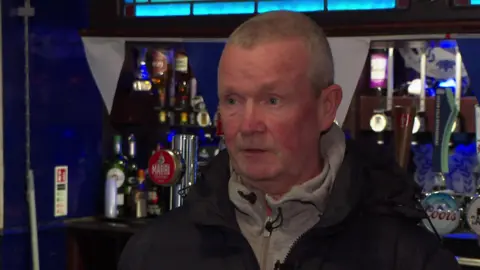 Donal Morgan, who has short grey hair and wears a black coat over a grey zip up hoodie, speaks to the camera in front of a bar lined with beer taps. 