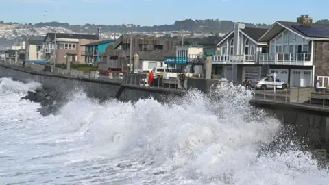 Waves hit the coastline in Pacifica, California. A couple of bystanders are looking at the waves and taking photos with houses lining the street behind them.