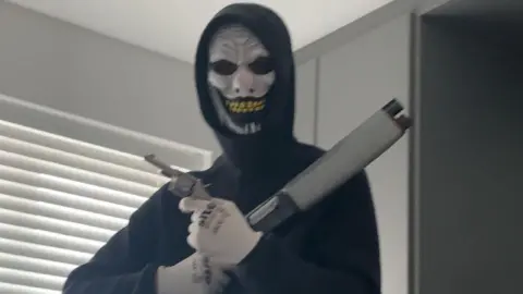 A masked man wearing a black hooded top and white gloves while holding a shotgun and a handgun. He is standing indoors in front of a window which is covered with blinds.