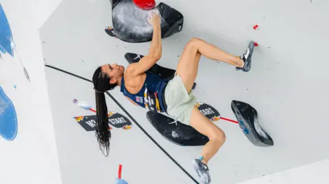 International Federation of Climbing/ Lena Drapella Molly Thompson-Smith climbing. Molly, in a Team GB vest and green shorts, clings to a climbing wall, one leg hanging loose. She has a determined look and her long ponytail hangs down. 