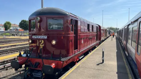 A burgandy coloured vintage train is pulled up alongside a platform with a number 12 written in gold colours and the desination of Amersham written on the front. 