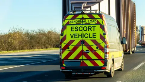 A red and yellow striped van marked with 'abnormal load - escort vehicle' drives behind a lorry bearing an oversized square load.