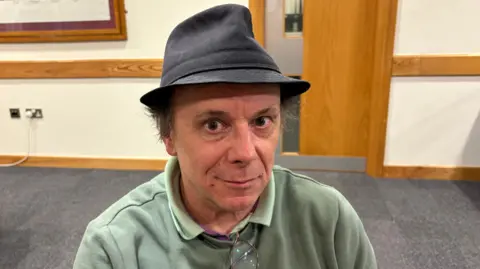 Martin Heath/BBC Ben Foley in his wheelchair wearing a grey trilby and green sweater. He is sitting in a council foyer with a grey carpet and white walls.