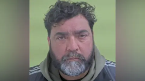 ISLE OF MAN CONSTABULARY Police photo of Samsha Sanghera, who has dark hair that is curly on top and a dark and grey beard. He is wearing a khaki hoodie under a tracksuit top.