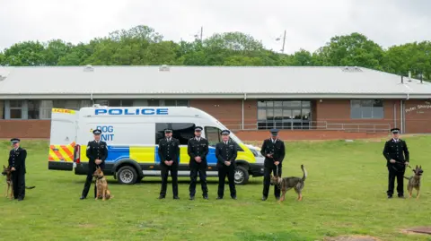 Hertfordshire Constabulary Seven male police officers, all standing outside, in front of a large brick building with a large metal roof. Four of the men are holding on to dogs. They are by a large police dog van. 