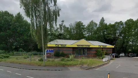 Google Maps The image shows the intended location for the new Camberley Burger King. There is a vacant yellow car wash built with red bricks. There are bushes and grass in front of the store with tall, green trees behind the shop. Some cars can be seen parked in the background on the right-hand-side. The sky is grey and cloudy.