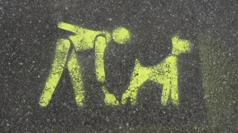 A sign on a road telling people to scoop up their dog poop when they take them out for a walk.