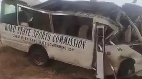 A badly damaged Kano State Sports Commission bus, with a crumpled front end and roof partially torn off