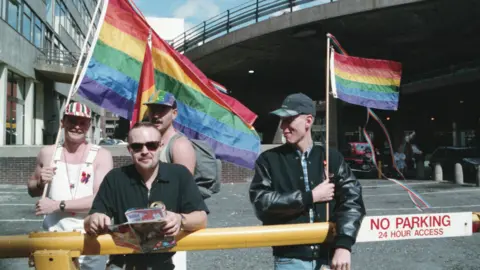 Stuart Rhoades Four men are standing at the side of a road in Manchester with colourful Pride flags.