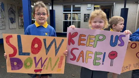 Two young girls stand outside a primary school. They are both dressed in a school uniform of a blue jumper and a yellow polo shirt underneath. The girl on the left holds a paper sign with the words 'Slow Down' written in colorful lettering. Beside her another girl holds a similar hand drawn sign with the words 'keep us safe" in bright bubble letters.