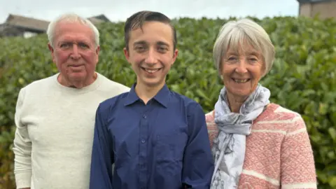 BBC Adrian, Daniel and Jill in a midshot in front of a tall hedge all smiling to camera.