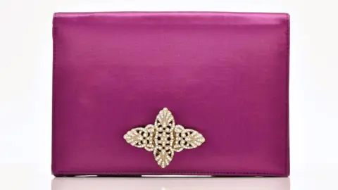 Halls A rectangular clutch bag that is silk and is a dark pink/mauve colour. It has an ornate brooch on the front that us gold with silver gems.