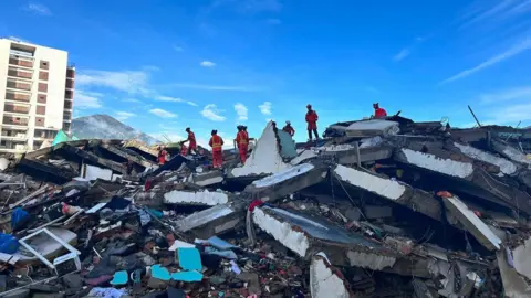 A group of about ten rescuers in orange uniforms and helmets are working on top of a pile of rubble with large concrete slabs and smaller items. Behind, a block of flats remains standing, and a mountain is visible in the background, set against a blue sky with light white cloud.