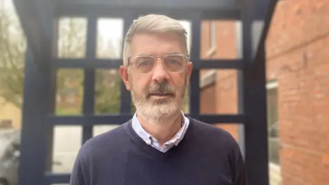 Tommy is standing outside. He's near a building. He wears glasses and has a navy jumper and a light coloured shirt on. A wood frame is behind him. A car is in the background. The background is blurred and Tommy is in focus.