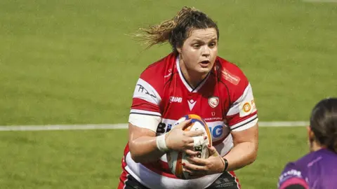 Gloucester-Hartpury back row Sarah Beckett in possession during a Premiership Women's Rugby match, wearing the red and white striped Gloucester home shirt.