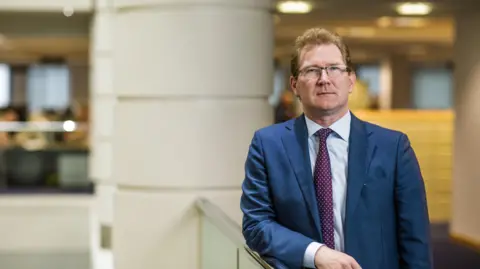 Government of Jersey A man with short, light-brown hair and glasses. He is wearing a light shirt and maroon tie with light-coloured dots, underneath a light blue blazer. He is leaning against a waist-height railing, inside what appears to be an office building. A circular white pillar is visible behind him. He is staring directly at the camera, to the right of the image.