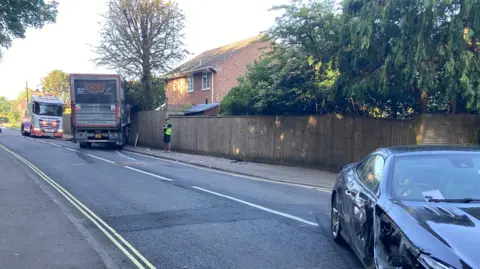 Lorry protruding from a garden fence with debris across the road a man in high-vis jacket is standing by the fence a black car with a crushed side is across the road.
