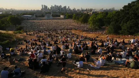 pA A large crowd sits on a parched hillside in a park, with the Canary Wharf skyline beyond.