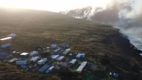 TERRES AUSTRALES ET ANTARCTIQUES FRANCAISES A coastline with large plumes of smoke in the distance. In the foreground there is a small collection of mobile buildings making up the La Roche Gordon research station.