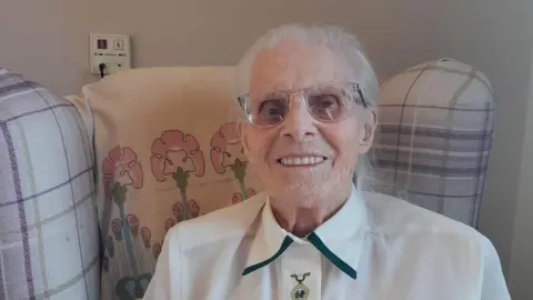 A woman sitting in a tartan armchair, smiling at the camera. She is wearing a white, collared blouse and clear-framed glasses. She has fine grey hair tied back.