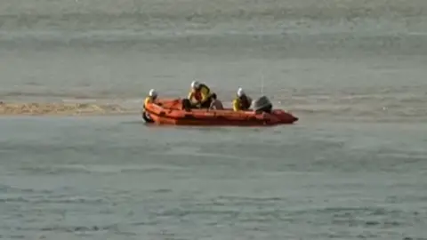 BBC The boy sitting onboard a small orange lifeboat, being helped by three crew members wearing yellow jackets, red life jackets and white helmets. The boat is surrounded by water, with the exception of a small bar of sand to the left.