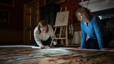 Mike Hodgson Two women looking at a Tudor tapestry which has been laid on the floor. Both women are kneeling down. One of them is wearing a beige jumper and another woman is wearing a blue knit jumper. They are using a torch to look at the tapestry.