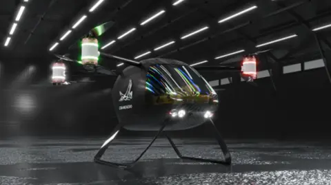 CraneAero A prototype vertical take-off and landing aircraft with four separate rotors on the ground in what looks like a large, dark empty warehouse. It has lights on the front and on each rotor. The aircraft looks like a helicopter but with four separate rotors instead of one big one.