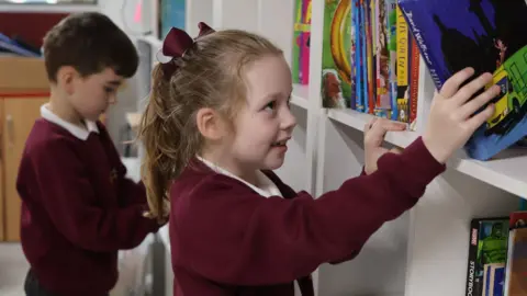 Blacklow Brow School A young girl with dark blonde hair, wearing a burgundy school cardigan and a red bow, selects a blue children's book from a large white bookshelf. A young boy, also in uniform, looks at a book in the backdrop. 