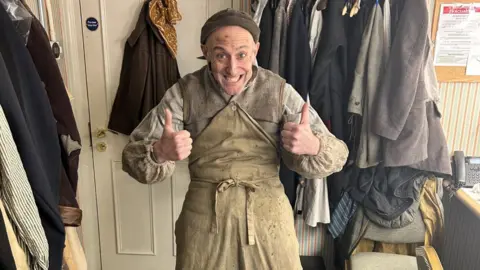 Jamie Tyler Jamie in a dressing room, with costumes all around him, wearing a dirty apron, a dirty striped shirt, a grey waistcoat and a hat. He has both his thumbs up and is smiling.
