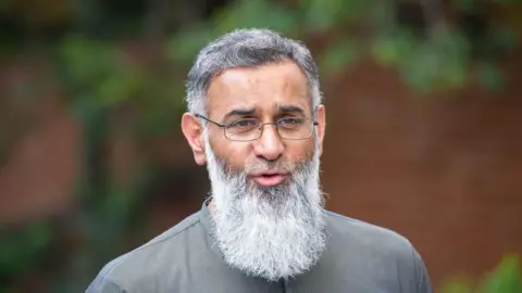 PA Anjem Choudary  pictured outside. He has glasses, greying hair and a large grey beard. It is a candid photo and he looks like he is about to speak.