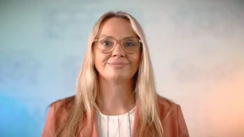 A photograph of a woman wearing glasses looking directly at the camera. She has blonde hair. 