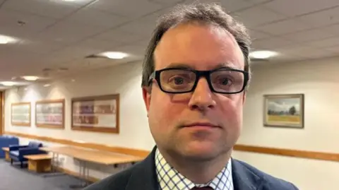 Martin Heath/BBC Henry Vann with short dark hair and black-framed glasses, looking at the camera while wearing a black jacket, white shirt with squared pattern and tie. He is standing in a carpeted corridor with paintings on the wall behind him and wooden tables and blue chairs lined up against the wall.